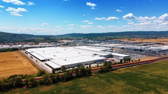 Large manufacturing plant surrounded by farmland. White industrial roofs and colorful railcars extend between green crops, harvested fields, and wooded hills.