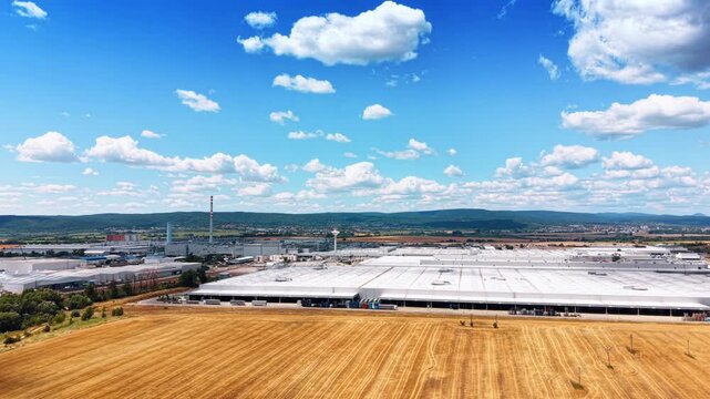 Wide aerial view of a massive production plant. An expansive white roofed factory building sits adjacent to a dry agricultural field under a cloudy sky.