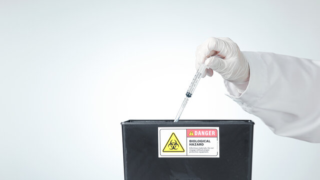 Cautious scientist hand wearing white glove drops syringe into black biohazard disposal bin inside laboratory during safe waste procedure