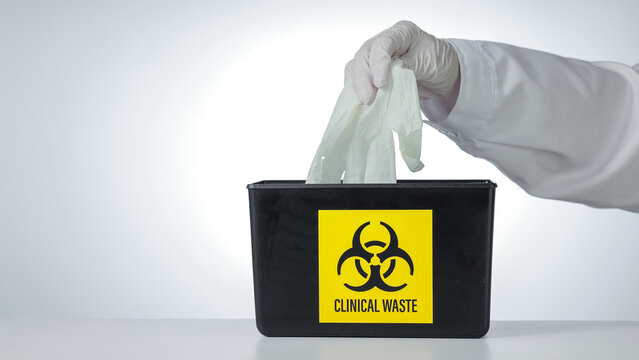 Cautious professional hand drops protective medical glove into black clinical waste biohazard disposal bin with yellow symbol