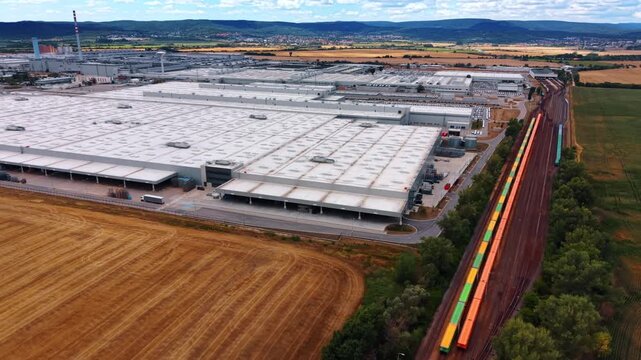 Aerial view of factory rooftops and striped chimne. A tall striped chimney rises above factory buildings beside a railway line bordered by golden and green farmland.