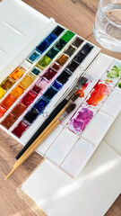 Fototapeta premium Watercolor Paint Palette with Brushes and Paper on Wooden Table