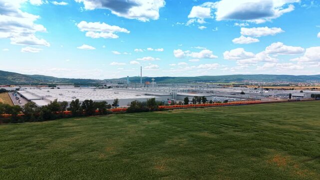 Grassy foreground overlooking automotive plant and railway. Green grassland stretches before a factory complex and freight train, bordered by hills and scattered clouds.