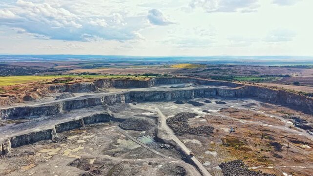 Granite quarry is visible with a large pit dug into the earth. Surrounding land is flat with fields and hills. Sky has some clouds and blue areas. No activity is seen in the quarry.