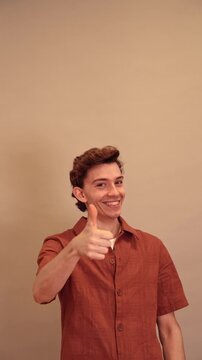 Happy young man giving thumbs up while pointing sideways on beige background