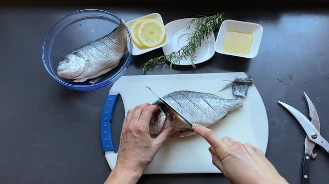 Top view of hands scoring fresh raw gilthead seabream fish with a knife on a cutting board, preparing for cooking with lemon, rosemary and olive oil on dark background.