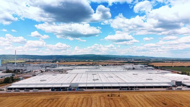 Manufacturing complex beneath large cumulus clouds. Wide white roofs, loading bays, golden stubble, distant farmland, and wooded hills span the aerial scene.