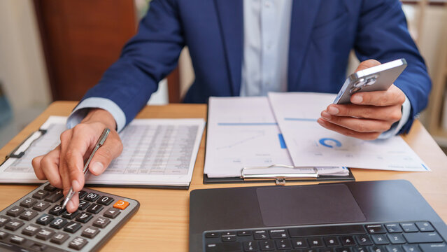 Businessman reviews financial reports with a calculator and smartphone, managing budgets, expenses, and accounting records for efficient business finance and informed decision-making.