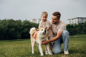Obraz premium Father and son interacting with a friendly dog in a green park, child sitting on father's knee while petting the dog's back, modern buildings visible in the background