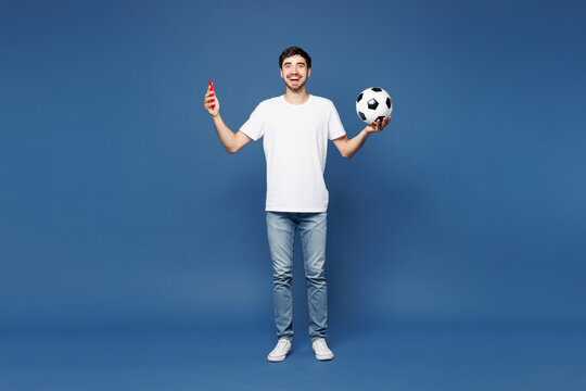 Full body young happy overjoyed man fan wears white t-shirt cheer up support football sport team hold in hand soccer ball watch tv live stream use mobile cell phone isolated on plain blue background.