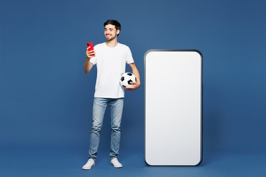 Full body young man wear white t-shirt cheer up support football sport team hold soccer ball watch tv live stream big blank screen mobile cell phone use smartphone isolated on plain blue background