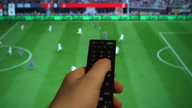 Hand holding TV remote control watching football match on screen.