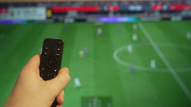 Hand holding TV remote control watching football match on screen.
