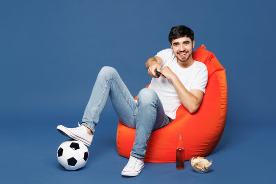 Full body young bored man fan wear white t-shirt cheer up support football sport team hold in hand soccer ball watch tv live stream sit in bag chair switch channel isolated on plain blue background