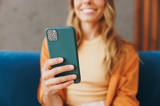 Cropped closeup young woman wear brown shirt hold in hand use mobile cell phone sit on blue sofa couch stay at home hotel flat rest relax spend free spare time in living room indoors. Focus on device.