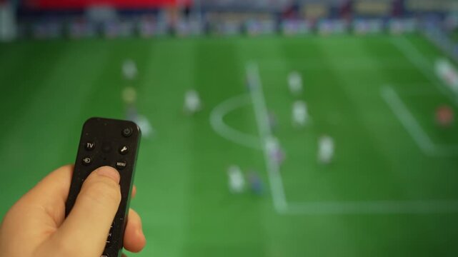 Hand holding TV remote control watching football match on screen.