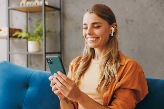 Young woman wear brown shirt listen music in earphones use mobile cell phone sit on blue sofa stay at home flat rest relax spend free spare time in living room indoors grey wall People lounge concept