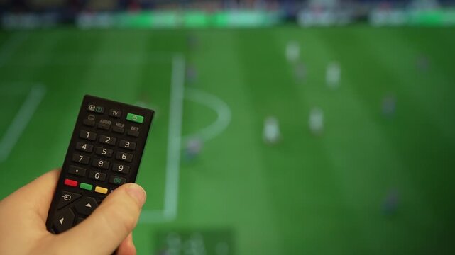 Hand holding TV remote control watching football match on screen.