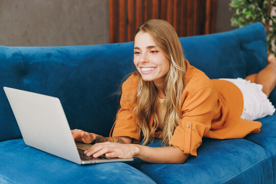 Smiling smart young IT woman wear brown shirt use work on laptop pc computer lay down blue sofa couch stay at home hotel flat rest spend free spare time in living room indoors. People lounge concept.
