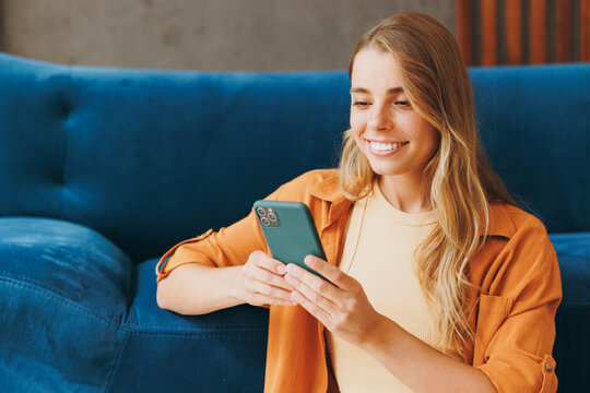 Young woman wear brown shirt hold in hand use mobile cell phone sit at blue sofa couch stay at home hotel flat rest relax spend free spare time in living room indoors grey wall. People lounge concept.
