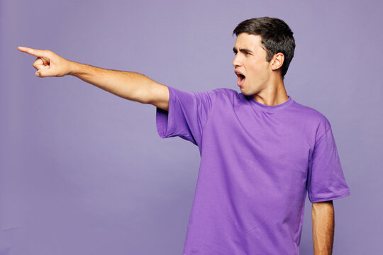 Young sad mad furious outraged Caucasian man he wear violet t-shirt casual clothes point index finger aside scream isolated on plain pastel light purple background studio portrait. Lifestyle concept.