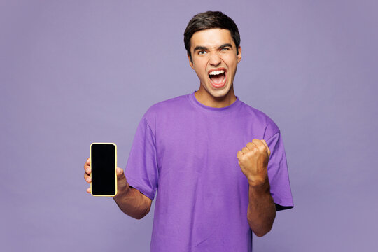 Young man wear violet t-shirt casual clothes hold in hand use mobile cell phone with blank screen workspace area do winner gesture isolated on plain pastel light purple background. Lifestyle concept.