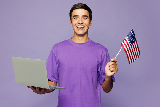 Young smart fun IT man he wearing violet t-shirt casual clothes hold American flag use work on laptop pc computer isolated on plain pastel light purple background studio portrait. Lifestyle concept.