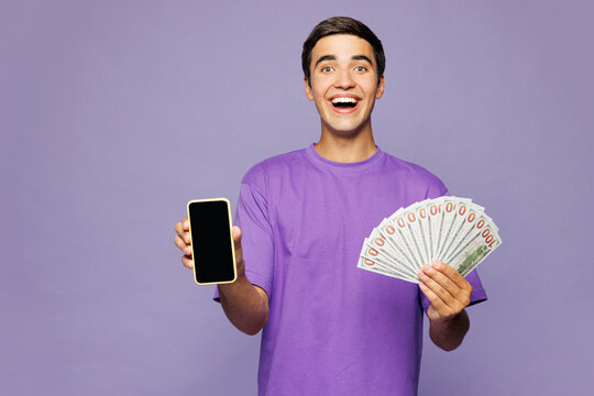 Young man wear violet t-shirt casual clothes use blank screen mobile cell phone hold in hand fan of cash money in dollar banknotes isolated on plain pastel light purple background. Lifestyle concept.