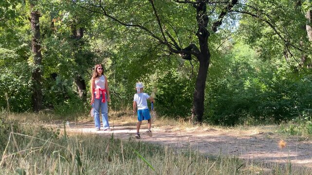 A mother and her young son play badminton together on a dirt path in a sunny forest. They share a playful, happy moment surrounded by green trees.