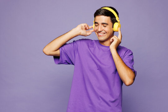 Young happy Caucasian man he wear violet t-shirt casual clothes listen to music in headphones raise up hands dance isolated on plain pastel light purple background studio portrait. Lifestyle concept.