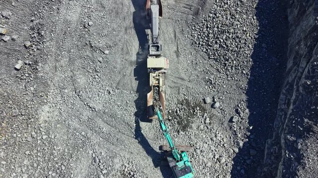 A large machine works in a granite quarry. It cuts and moves stone and materials. Workers oversee the operation in a busy mining area. Rocks and dust fill the space.