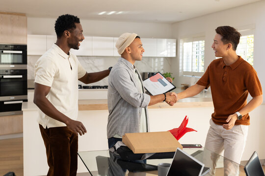 Diverse male coworkers shaking hands holding tablet by laptop gift box on glass table in kitchen