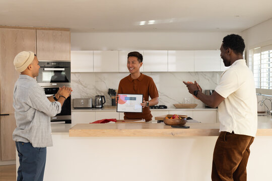 Diverse men in polos clapping, center Asian man holding document at kitchen island with fruit bowl