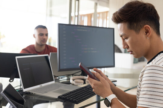 Diverse male coworkers using smartphone and coding on laptop with external monitor showing code