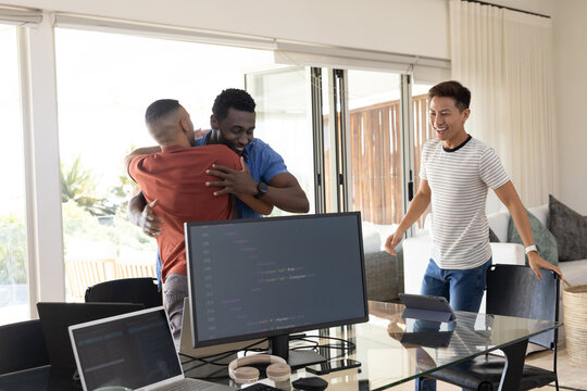 Diverse male coworkers hugging near glass dining table at home with monitor showing code