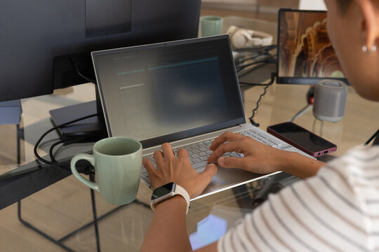 Mid-adult Asian man typing on slim laptop at glass-top desk, striped shirt, green mug, copy space