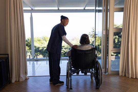 African American nurse in scrubs placing hand on senior wheelchair, gazing at water via glass doors