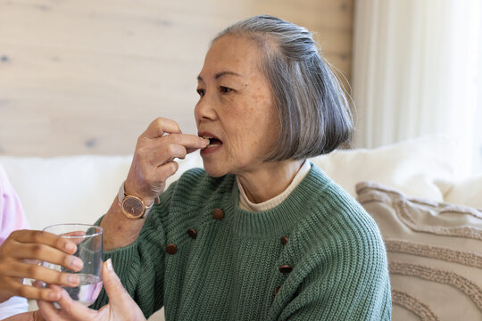 Senior Asian woman in green sweater with watch taking pill on sofa, caregiver holding glass