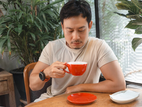 Thoughtful Asian man sips coffee from a red mug, relaxing in a cozy indoor setting.