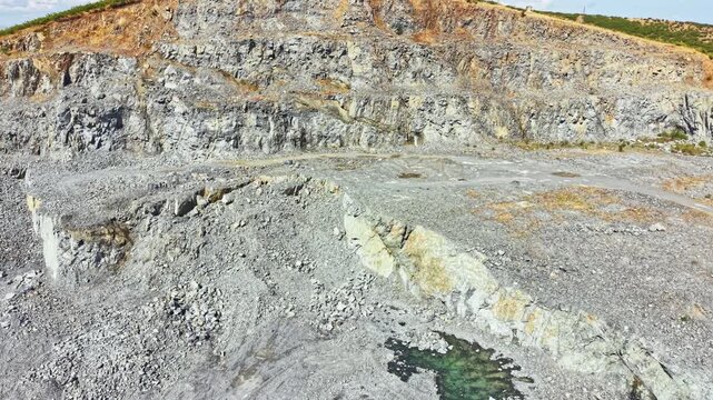 The granite quarry reveals steep rock walls and a large area of crushed stone. Heavy machinery is seen working in the pit near water pools. The sky is clear and bright.