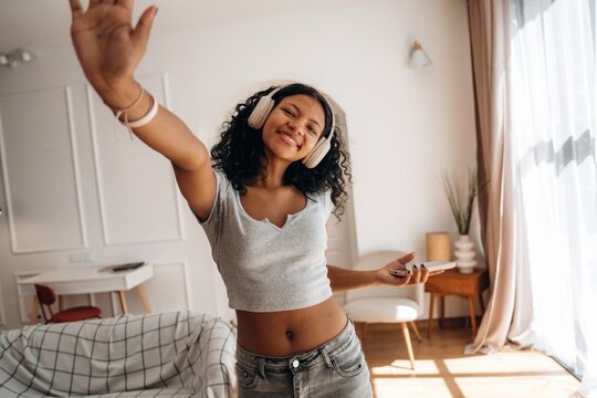 Young woman with curly hair wearing headphones smiles and waves while holding a smartphone in a bright living room with modern decor and natural light