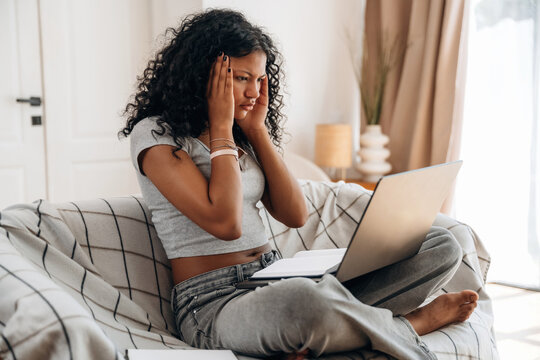 Young woman with curly hair sitting on a couch, looking stressed while working on a laptop in a cozy living room with soft lighting and decorative elements