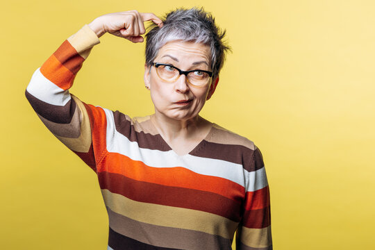 Mature woman looking confused and thoughtful, scratching her head against a yellow background, experiencing memory issues