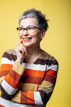 Senior woman with short gray hair and glasses smiling thoughtfully, touching her chin against a vibrant yellow background