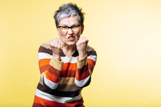 Senior woman showing intense anger and frustration, clenching fists on a yellow background, embodying feelings of conflict and rage