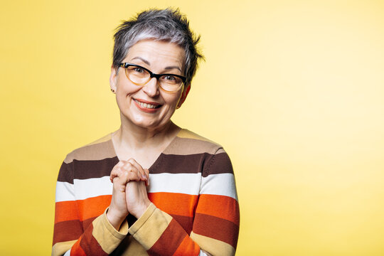 Senior woman with gray hair and glasses, holding hands, smiling warmly and looking at the camera, showing gratitude