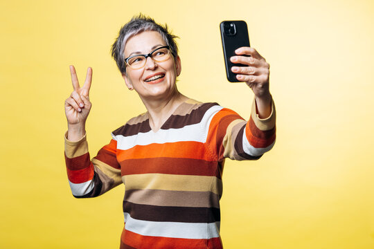 Smiling woman with short grey hair and glasses taking a selfie on her smartphone, showing a peace gesture