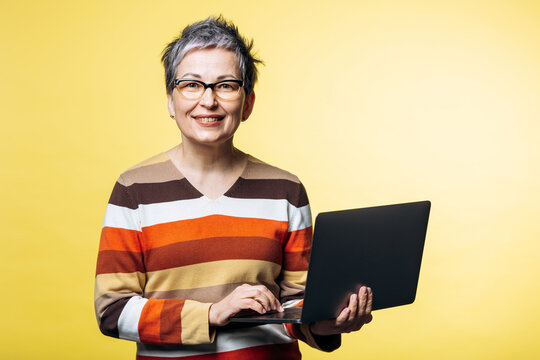 Smiling senior woman wearing eyeglasses and a striped sweater, engaging with a laptop computer on a yellow background