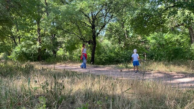 A mother walks along a dirt path toward her young son holding a racket. They enjoy a peaceful, joyful walk in a sunny green forest.