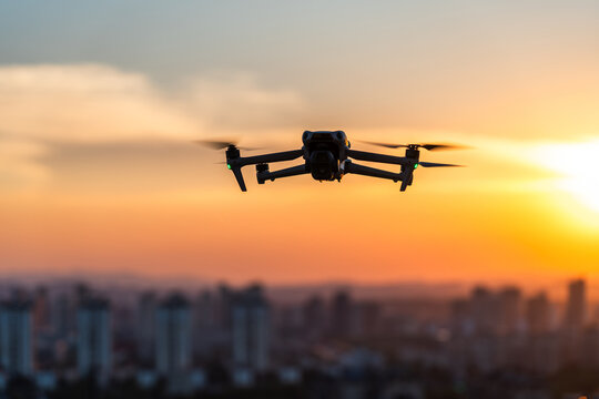 A quadcopter drone flies in silhouette against a sunset sky above a blurred urban cityscape during evening hours.